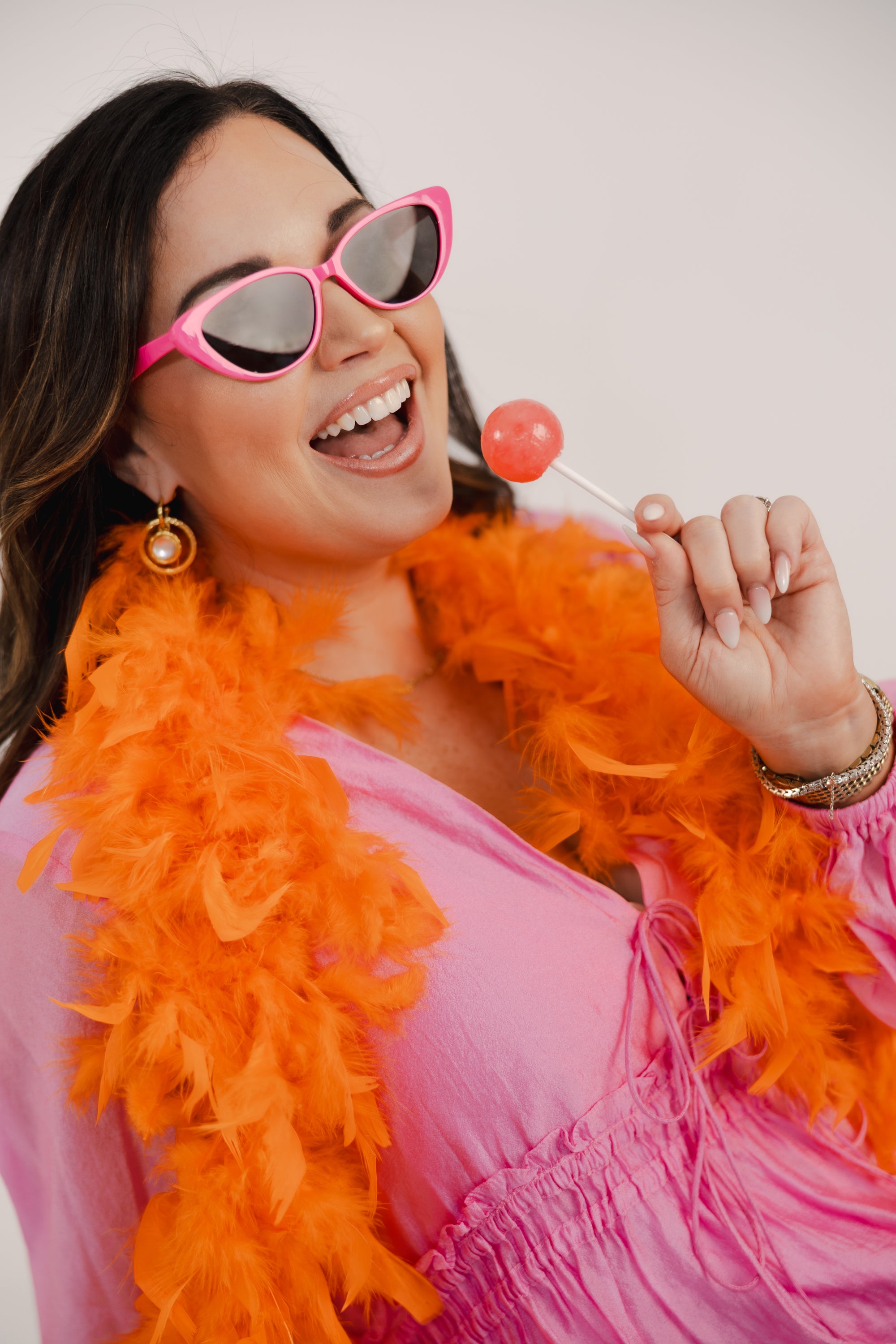 Woman wearing pink sunglasses and an orange feather boa, holding a lollipop.
