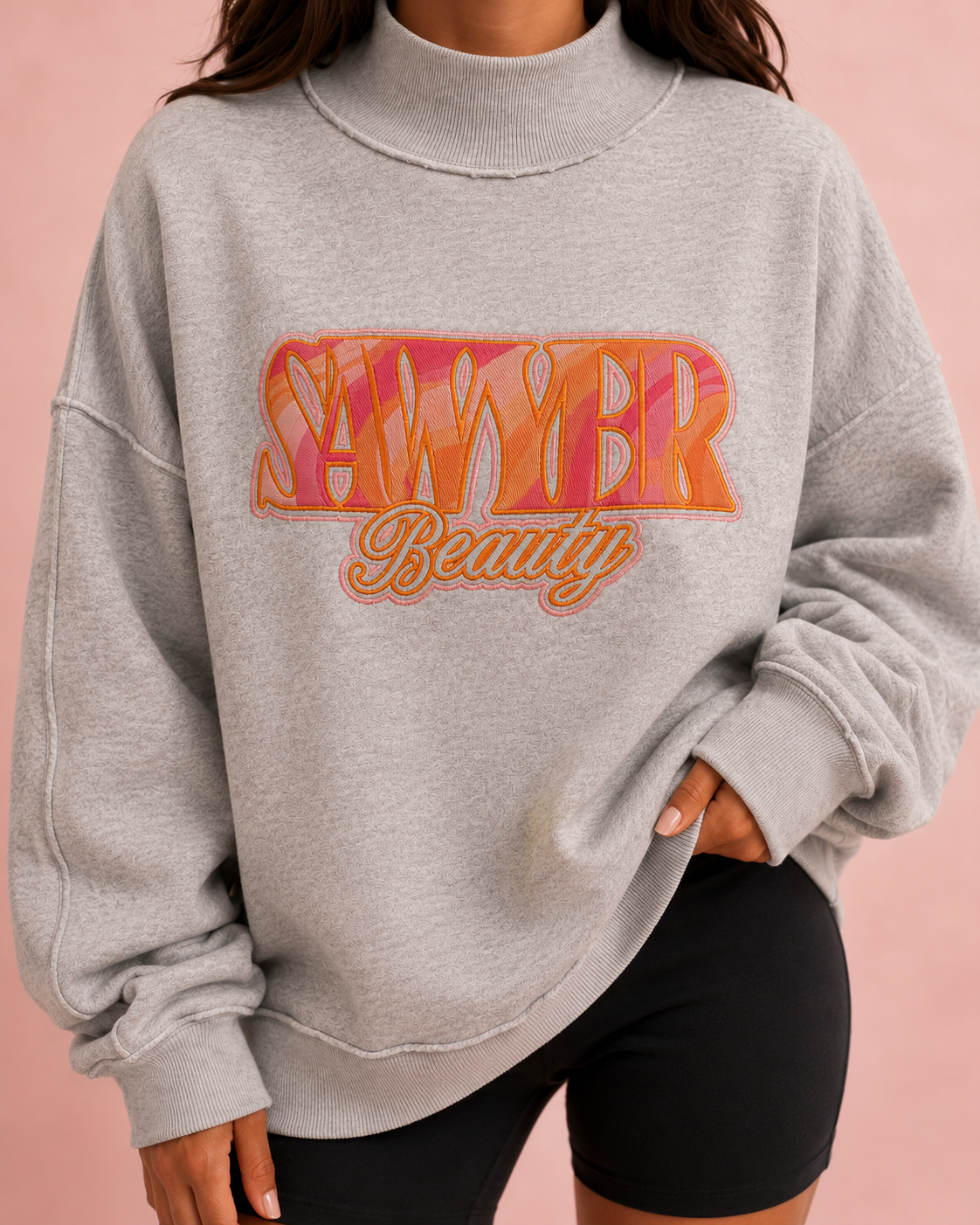 A person wearing an oversized light heather gray mock-neck sweatshirt with a colorful 'Sawyer Beauty' graphic on the front, paired with black shorts, against a solid pink background.