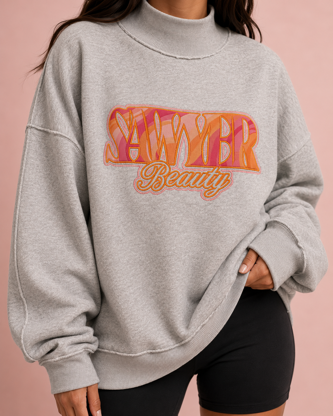 A woman wearing a light gray heather crewneck sweatshirt with a colorful 'SAWYER Beauty' graphic on the front, paired with black shorts, set against a solid pink background.